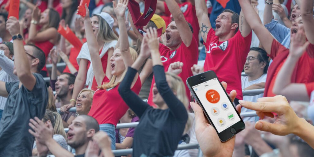 A person holding a mobile phone about to trigger a Rave Alert while cheering fans enjoy the game in the background.