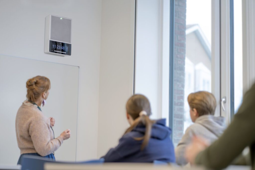 A classroom of students and their teacher look at the Axis C1710 Network Display Speaker hanging on the wall.