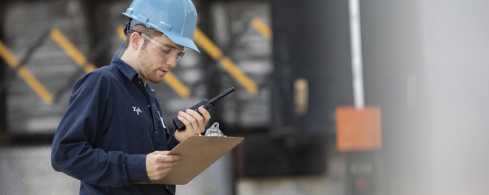 A logistics worker uses a two-way radio and a clipboard while examining a warehouse workflow.